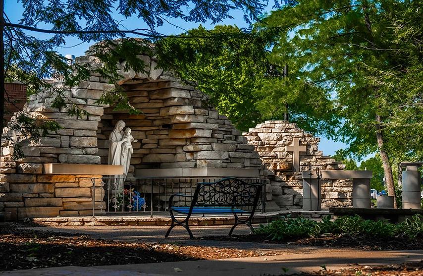 A bench under a tree faces a stone wall and statue.